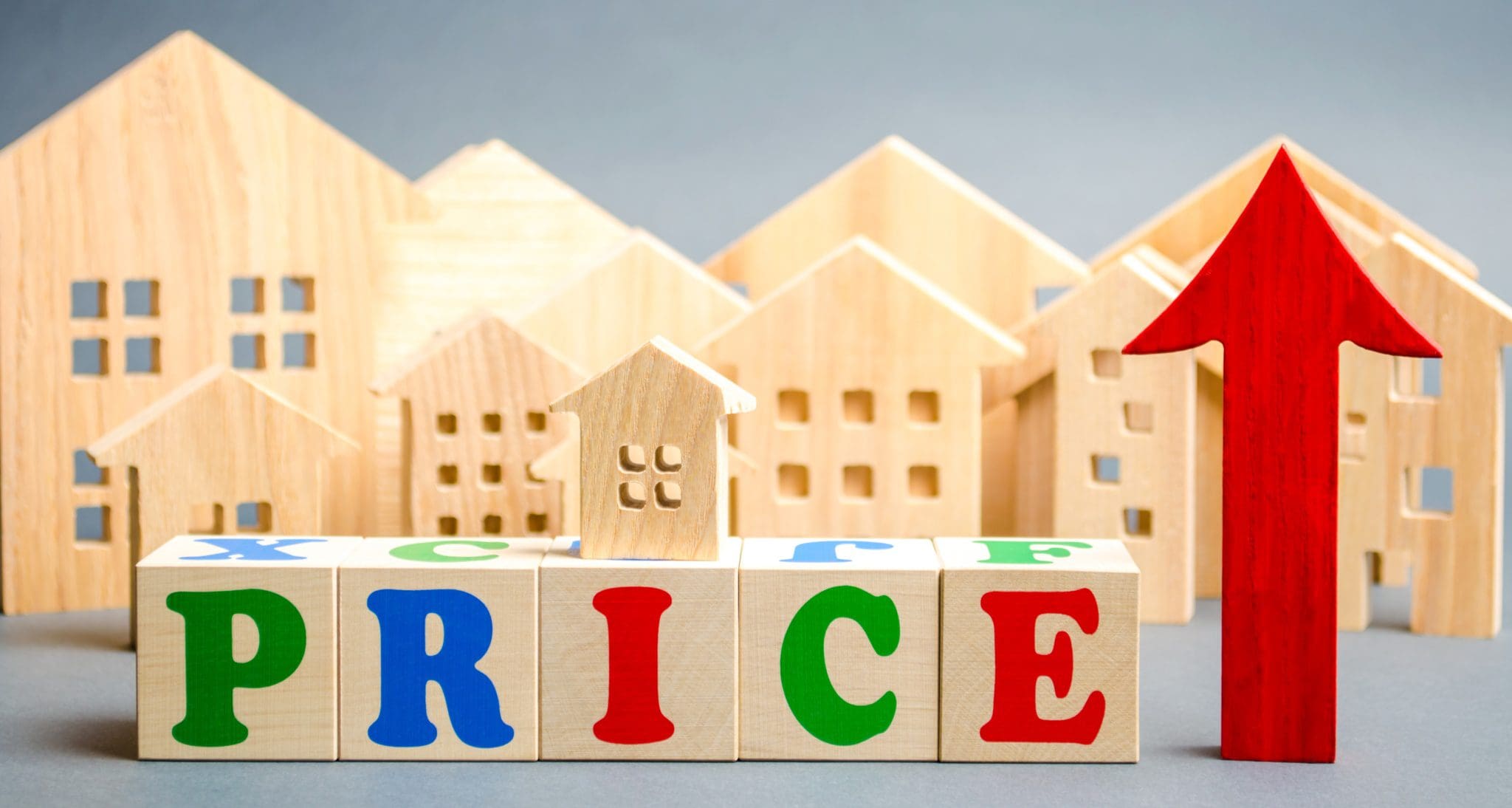 Wooden toy houses in the background with colorful alphabet blocks spelling “PRICE” in the foreground, a small house sitting on the blocks, and a large red upward arrow symbolizing rising property prices in the housing market.